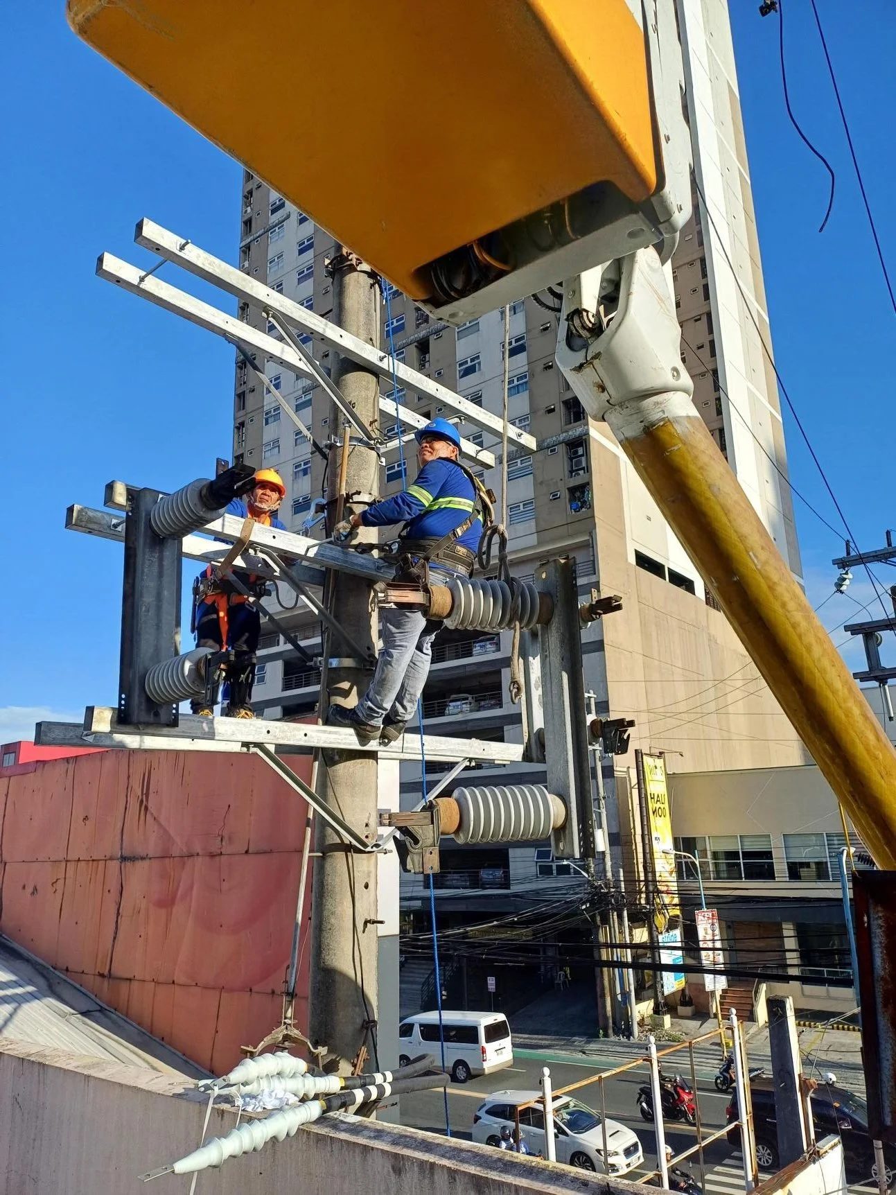 Two utility workers wearing safety gear working on an electrical pole, surrounded by urban buildings and street traffic.