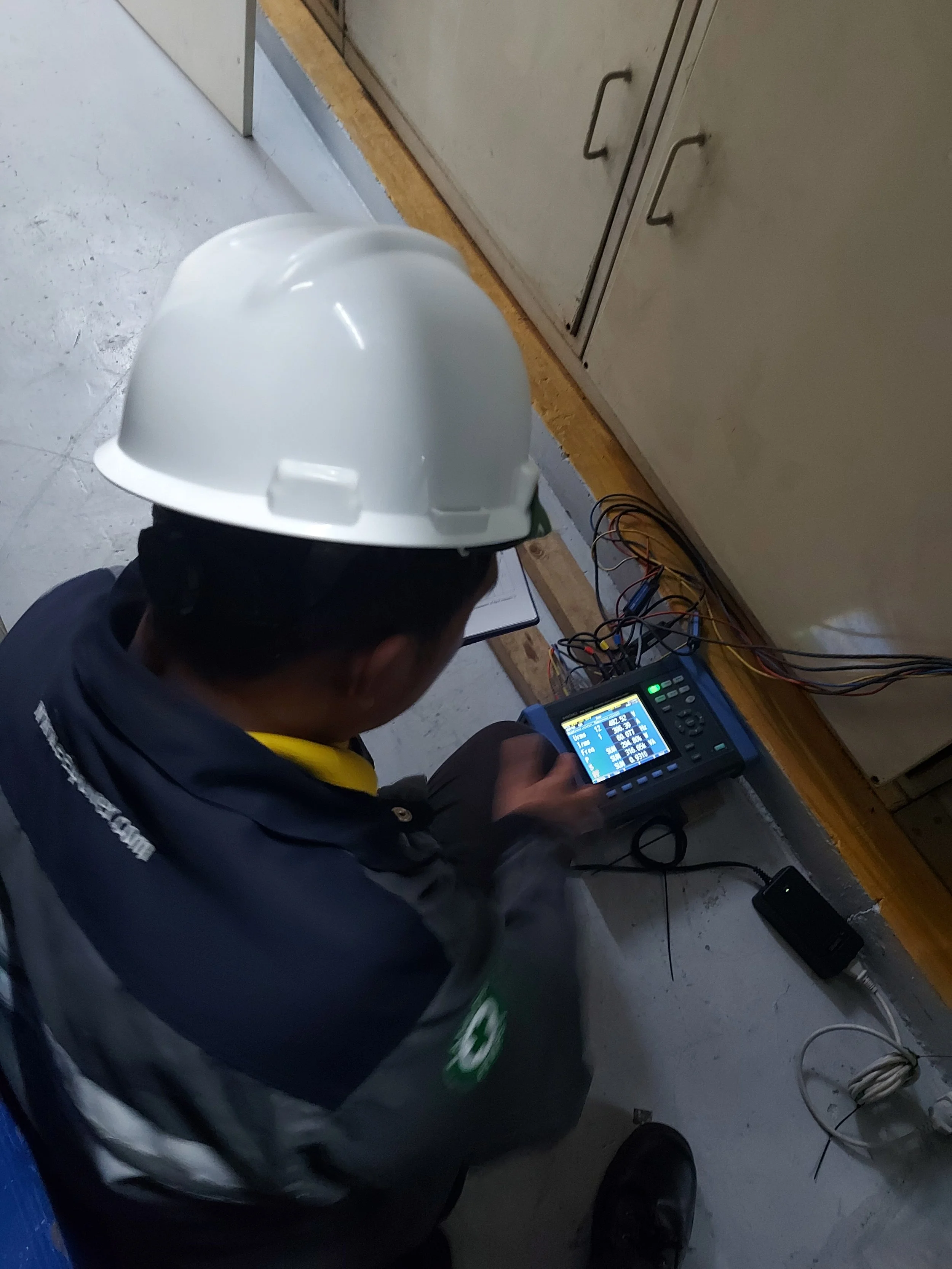 A construction worker wearing a white safety helmet sitting on the floor and operating a testing device connected to multiple wires, at a construction site.