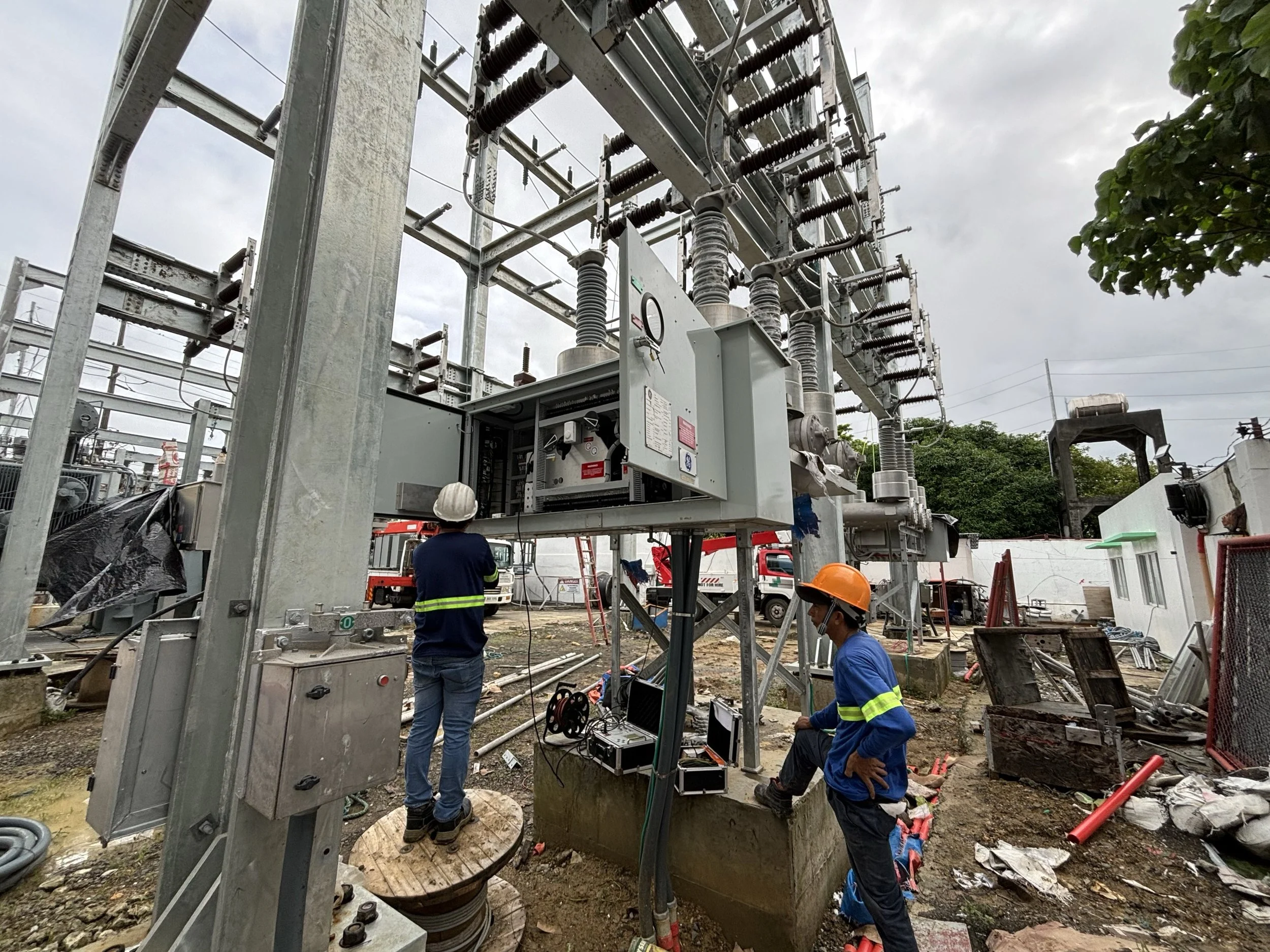 Two construction workers in hard hats working on electrical equipment at an outdoor construction site with workers and equipment visible in the background.