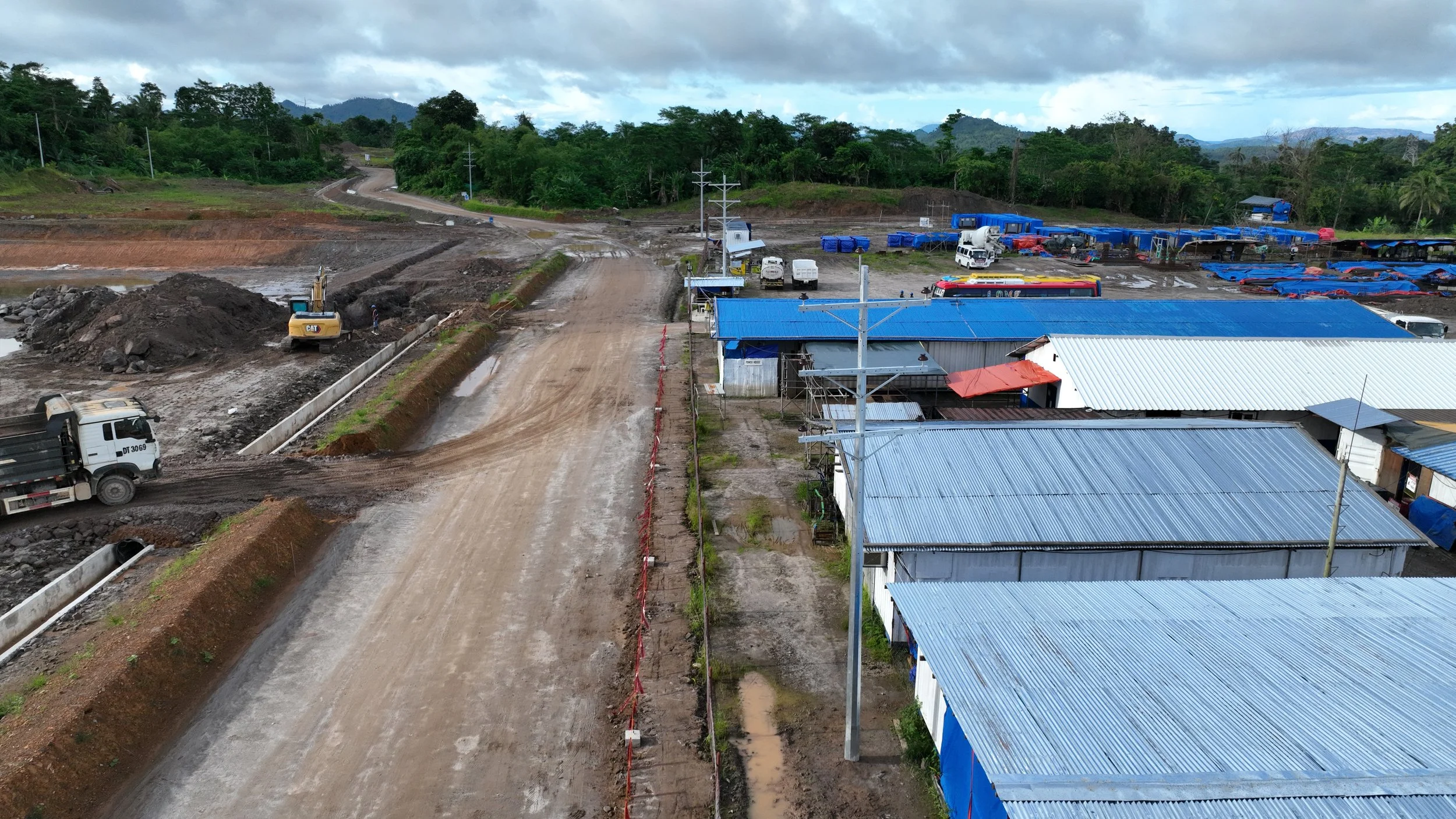 Construction site with dirt road, construction vehicles, and buildings with metal roofs, surrounded by greenery and trees, under cloudy sky.